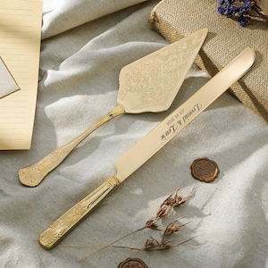 Puede incluir: Juego de servidor y cuchillo para pastel de color dorado. El servidor tiene un diseño floral ornamentado en la hoja. El cuchillo está grabado con "Leonid & Laurie 03.15.2026". El conjunto se muestra sobre una superficie neutra, temática de boda.