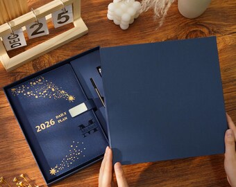 May include: A navy blue 2026 daily plan journal with gold starburst designs, presented in a matching gift box. The journal includes a pen and a silver clasp. The name "Michael" is printed on the journal. A wooden calendar is in the background.