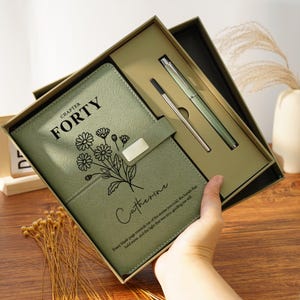 Puede incluir: Un juego de diario y bolígrafo verde salvia en una caja de regalo. La portada del diario presenta el texto "Chapter Forty", un diseño floral y el nombre "Catherine". Se incluye un bolígrafo y una recarga plateados.
