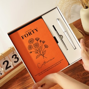 Può includere: Un diario arancione con il testo "CAPITOLO QUARANTA" e un'illustrazione floreale, presentato in una confezione regalo bianca con una penna bianca. Il diario ha anche il nome "Catherine" e una citazione.