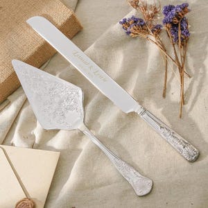 May include: A silver cake knife and server set with ornate embossed floral patterns on the handles and server blade. The knife blade is engraved with the text Leonid & Louie and the date 05.18.2024 in a script font, featuring a serrated edge.