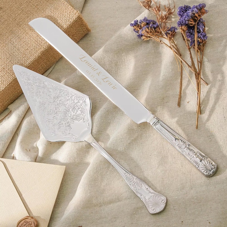 May include: A silver cake knife and server set with ornate embossed floral patterns on the handles and server blade. The knife blade is engraved with the text Leonid & Louie and the date 05.18.2024 in a script font, featuring a serrated edge.