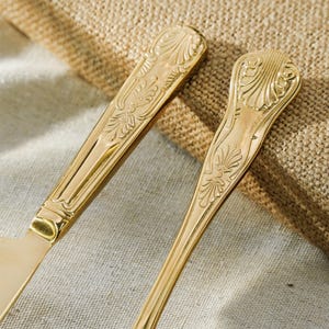 May include: Two gold-toned metal utensils featuring ornate, embossed shell and leaf patterns on the handles. The polished finish reflects light, highlighting the intricate decorative details carved into the surface of the flatware handles.