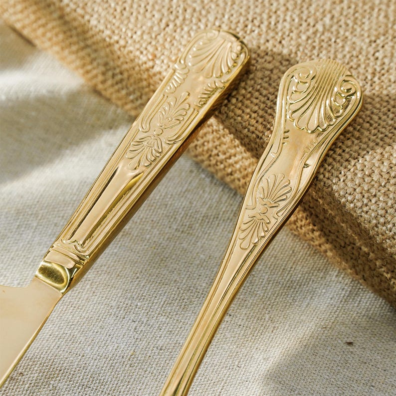 May include: Two gold-toned metal utensils featuring ornate, embossed shell and leaf patterns on the handles. The polished finish reflects light, highlighting the intricate decorative details carved into the surface of the flatware handles.