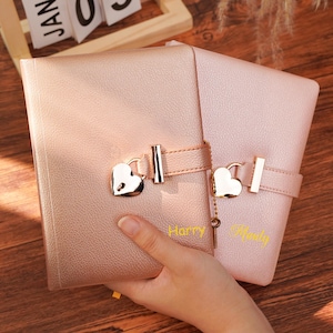 May include: Two leather-bound journals, one rose gold and one light pink, are displayed. Each journal features a heart-shaped lock and a strap closure. The rose gold journal has the name "Harry" in gold lettering, and the pink journal has the name "Mandy".