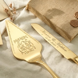 May include: A gold-coloured wedding cake server and knife set. The server has an ornate floral design and the knife is engraved with the names "Monroe & Matthew" and the date "12-07-2025". The set is displayed on a white cloth.