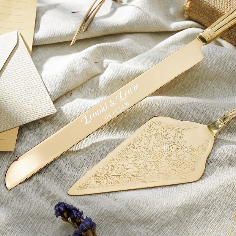 May include: A gold-toned cake knife and server set. The knife blade features the engraved text "Leonid & Lewie" and "03.15.2020". The triangular server blade is decorated with an intricate, embossed floral scrollwork pattern across its surface.