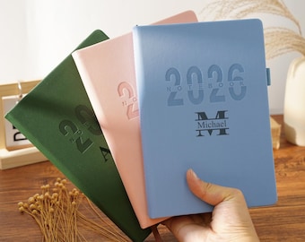 Op de afbeelding: Een hand houdt een lichtblauw notitieboek vast met de in reliëf aangebrachte tekst "2026 NOTEBOOK Michael". Twee andere notitieboeken zijn zichtbaar: één in roze en één in groen. Alle notitieboeken hebben een gestructureerde kaft en een lint als bladwijzer. De notitieboeken liggen op een houten oppervlak.