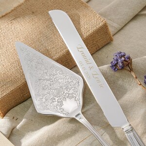 May include: A silver cake server with an intricate floral engraving and a matching silver cake knife featuring the engraved text "Leonid & Lenie" and the date "03.15.2026" on the blade, accompanied by a small sprig of dried purple flowers.