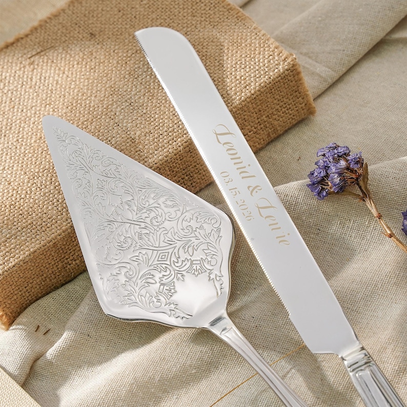 May include: A silver cake server with an intricate floral engraving and a matching silver cake knife featuring the engraved text "Leonid & Lenie" and the date "03.15.2026" on the blade, accompanied by a small sprig of dried purple flowers.