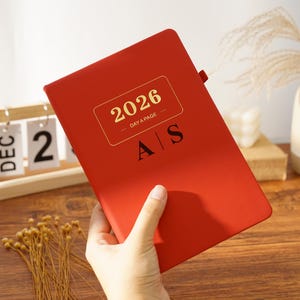 May include: A red 2026 day-a-page planner with gold lettering and the initials "A S". The planner is held by a hand and has a ribbon bookmark. The cover features the year 2026 in a gold rectangle.