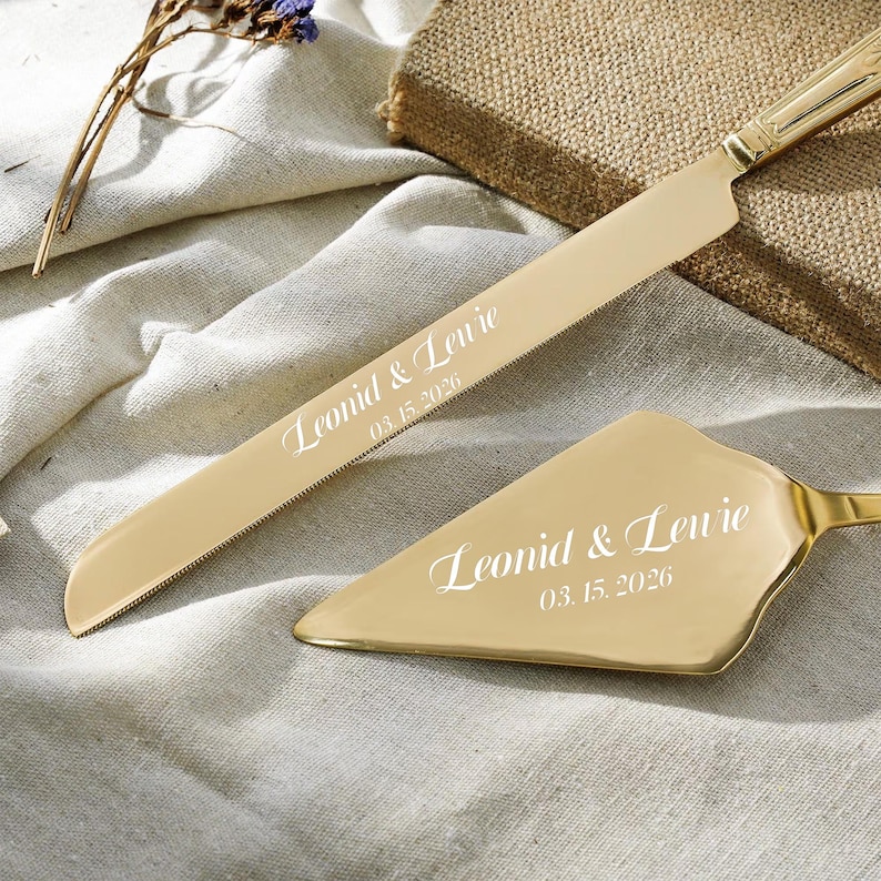 May include: A gold-toned cake knife and server set featuring a serrated blade and triangular head. Both pieces are engraved with the names Leonid & Lewie and the date 03.15.2026 in a white script font, showcasing a polished metallic finish.