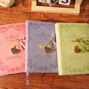 May include: Three journals in pink, lavender, and green, each with a heart-shaped lock and key. The journals have floral embossed designs and ribbon bookmarks. The word "Free" is visible on the lavender journal.