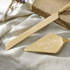 May include: A gold-toned cake knife and server set featuring a serrated blade and triangular head. Both pieces are engraved with the text Ashley & Christian and the date 03.15.2026 in a white script font, showcasing a polished metallic finish.