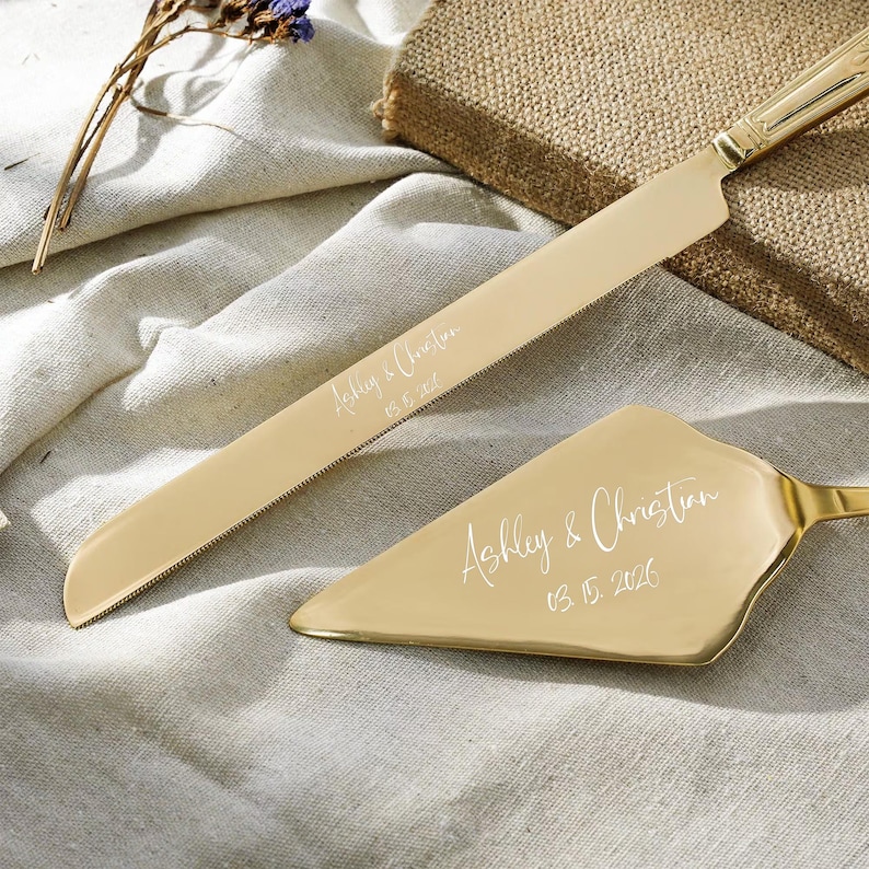 May include: A gold-toned cake knife and server set featuring a serrated blade and triangular head. Both pieces are engraved with the text Ashley & Christian and the date 03.15.2026 in a white script font, showcasing a polished metallic finish.
