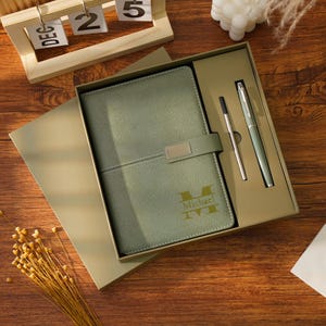 Può includere: Un set di diario e penna verde salvia in una confezione regalo. Il diario ha una chiusura a cinturino con un accento argentato e il nome "Michael" inciso sulla copertina. Sono inclusi una penna e una ricarica argentate.