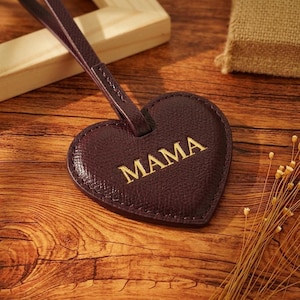 May include: A heart-shaped dark burgundy luggage tag with a textured surface and a matching strap. The word MAMA is embossed in gold capital letters across the centre of the heart. The tag features visible stitching along the outer edge.