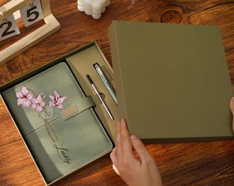 May include: A green journal with a floral design and the word "Lucky" is displayed in an open gift box. Two pens are also visible. The box is olive green. The journal has a magnetic closure. The background is a wooden surface.