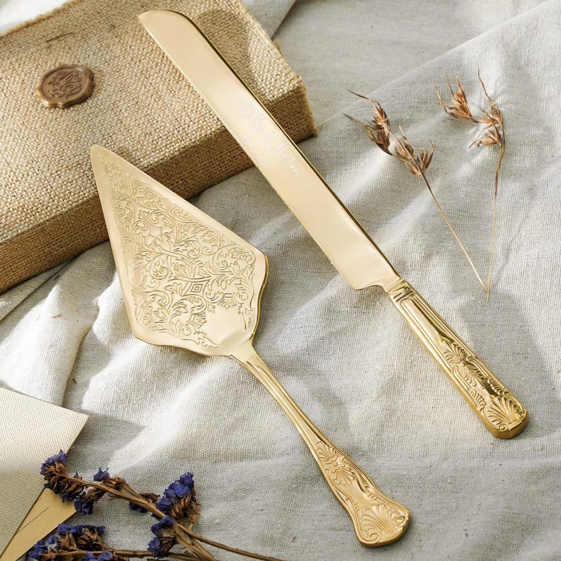 May include: A gold-toned cake serving set consisting of a triangular cake server and a long serrated knife. Both pieces feature intricate embossed floral and scrollwork patterns on the handles and the server blade, with a polished finish.