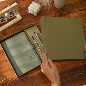 Op de afbeelding: Een saliegroen notitieboek- en pennenset gepresenteerd in een bijpassende geschenkdoos. Het notitieboek heeft een zilveren sluiting en de tekst "Sophia Conner". Een zilveren pen en een zwarte pen zijn inbegrepen. De doos wordt geopend.