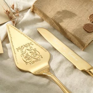 May include: A gold-toned cake server and knife set. The triangular server features an ornate engraved scrollwork pattern. The knife blade is engraved with the text "Monroe & Matthew" and a date. Both pieces have a polished, reflective metallic finish.