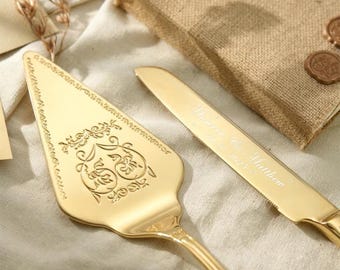 May include: A gold-toned cake server and knife set. The triangular server features an ornate engraved scrollwork pattern. The knife blade is engraved with the text "Monroe & Matthew" and a date. Both pieces have a polished, reflective metallic finish.