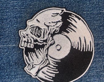 Op de afbeelding: Geborduurde patch met een schedelontwerp geïntegreerd in een vinylplaat. De schedel is wit met gedetailleerde schaduwen, de plaat is zwart en wit. Geschikt voor kleding of tassen.