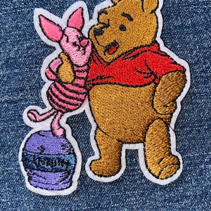 May include: An embroidered patch featuring a golden-brown bear wearing a red shirt standing next to a small pink piglet. The piglet stands atop a purple barrel with a blue band that has the word HUNNY embroidered in black thread on the front.