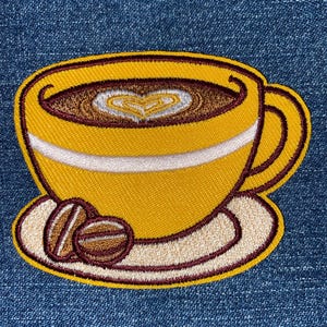May include: A yellow embroidered coffee cup with a brown heart design in the foam, a white rim, and a brown saucer with two brown coffee beans.