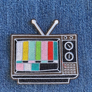 May include: Embroidered patch featuring a retro television set with a colorful test pattern on a blue denim background. The TV has a brown frame, white antenna, and black details. A fun, nostalgic accessory.