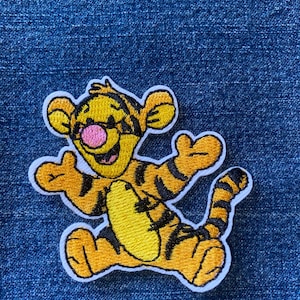 May include: An embroidered patch featuring a yellow and orange tiger character with black stripes, a pink nose, and a wide smile. The character is depicted in a joyful pose with arms raised, outlined in white thread against a blue denim background.