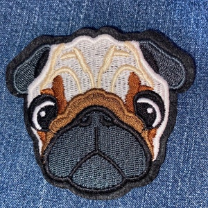 May include: Embroidered pug face patch with black, white, tan, and brown details. The patch features a detailed design of a pug's face, including its eyes, nose, and wrinkles. The patch is set against a blue denim background.