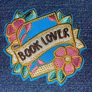 May include: Embroidered patch with the words "BOOK LOVER" on a banner. The patch features a blue background, pink flowers, and green leaves, all outlined in gold. The patch is on a denim background.