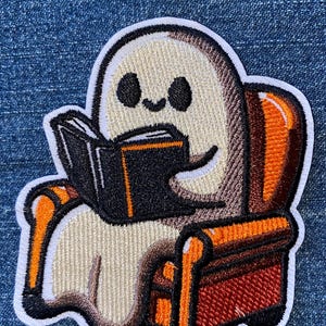 May include: An embroidered patch featuring a cream-colored ghost with black eyes and a small smile, sitting in a brown and orange armchair while holding an open black book with orange page edges. The design has a thick white border outline.