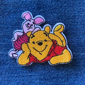 May include: An embroidered patch featuring a yellow bear wearing a red shirt lying down, with a small pink pig character standing behind the bear. The design is outlined in white thread and features detailed stitching throughout the characters.