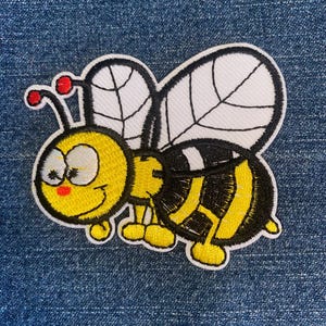May include: Embroidered patch featuring a cartoon bee with a smiling face. The bee has a yellow body with black stripes, white wings with black veins, and red antennae. The patch is on a blue denim background.