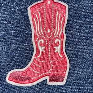 May include: A pink embroidered cowboy boot patch with white decorative stitching and a white border. The boot features a pointed toe and a small heel. The patch is set against a blue denim background.