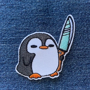 May include: Embroidered patch featuring a cartoon penguin holding a knife. The penguin has a black and white body with an orange beak and feet. The knife has a teal blade and a brown handle. The patch is on a blue denim background.