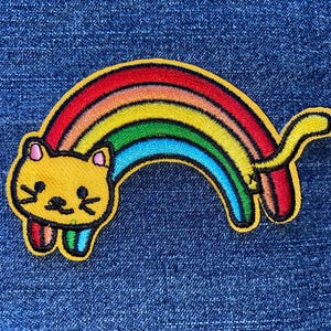 May include: A colorful embroidered patch featuring a yellow cat with a rainbow arch. The rainbow displays red, orange, yellow, green, and blue hues. The patch is set against a blue denim background.