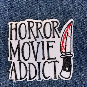 May include: Embroidered patch with the words "HORROR MOVIE ADDICT" in black script on a white background. A knife with a red, blood-like design is on the right. The patch is on a blue denim background.