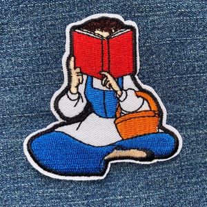 May include: Embroidered patch featuring a cartoon character reading a red book. The character is wearing a blue dress and white sleeves, with an orange basket. The patch has a white border and is set against a denim background.