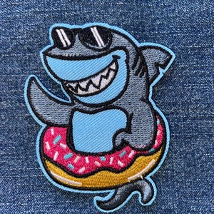 May include: Embroidered patch featuring a cartoon shark wearing sunglasses and floating on a donut-shaped float. The shark is gray and blue with a big smile. The donut is pink with sprinkles and a yellow outline.