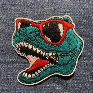 May include: Embroidered patch featuring a teal dinosaur head wearing red sunglasses. The dinosaur has a wide open mouth with white teeth and a red tongue. The patch is outlined in white and set against a blue denim background.