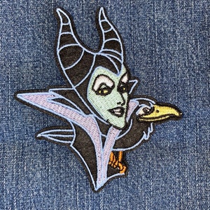 May include: Embroidered patch featuring Maleficent from Sleeping Beauty. The patch shows Maleficent's face with green skin, black horns, and a raven. The patch has a light blue outline and is set against a blue denim background.