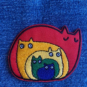 May include: A colorful embroidered patch featuring a family of cats nested inside each other. The patch has a red border, with a yellow cat, a green cat, and a blue cat inside. The patch is on a blue denim background.