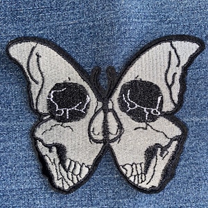 May include: Embroidered patch in the shape of a butterfly, with a skull design. The butterfly has gray wings with black outlines and details. The skull's eye sockets and teeth are black. The patch is on a blue denim background.