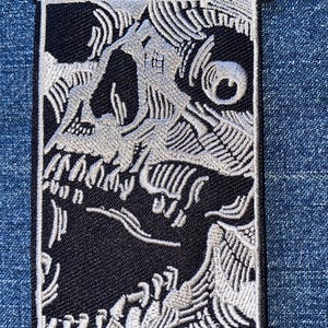 May include: A rectangular embroidered patch featuring a high-contrast black and white graphic design of a stylized skull. The intricate stitching creates detailed line work and shading, depicting the skull within a thin, rectangular border frame.