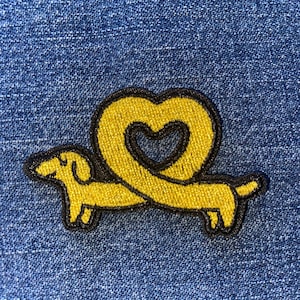 May include: Yellow dachshund dog patch with a heart-shaped tail, outlined in black, on a blue denim background. The patch features a playful design, perfect for adding a touch of whimsy to clothing or accessories.