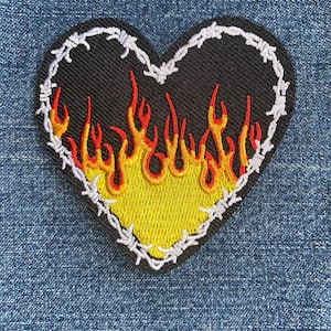 May include: Embroidered heart-shaped patch with a black background, yellow and red flames, and a white barbed wire border. The patch is likely designed to be sewn or ironed onto clothing or accessories.