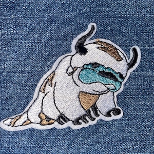 May include: White and brown embroidered patch of a cartoon animal with blue horns and a teal face. The animal is sitting on its haunches with its head down.